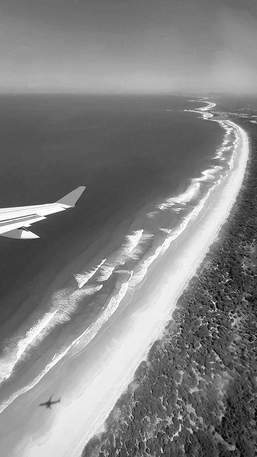 無料のおしゃれな縦長画像：飛行機の翼越しに広がる青い海と長く続く白い砂浜の空撮（白黒版）